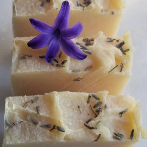 May include: Three bars of handmade soap with a light beige color and lavender buds. A purple flower is placed on top of the middle bar.