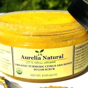 May include: A jar of Aurelia Natural organic turmeric citrus and honey sugar scrub. The jar contains a bright yellow scrub and has a black lid. The label reads "Net Wt. 227 g" and lists ingredients.