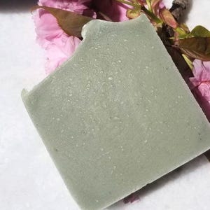May include: A square, pale green bar of soap with a textured surface. Pink cherry blossoms and a brown twig are in the background. The soap appears handmade and has a natural aesthetic.