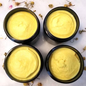 May include: Four open black jars filled with a smooth, yellow cream. The cream has a slightly textured surface. Dried flowers and petals are scattered around the jars on a white surface.