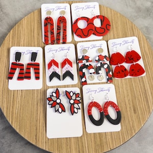 May include: A collection of nine pairs of earrings displayed on white cards labeled Stunning Statements. The earrings feature various geometric shapes in red, black, white, and silver, including arches, circles, chevrons, and floral-inspired designs.