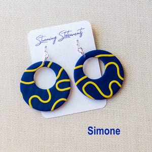 K&ouml;nnte beinhalten: Ein Paar kreisf&ouml;rmige Ohrh&auml;nger mit dunkelblauer Basis und gelbem Wellenlinienmuster. Die Ohrringe sind an einer wei&szlig;en Pr&auml;sentationskarte befestigt, auf der der Text Stunning Statements und Simone in blauer Schrift aufgedruckt ist.