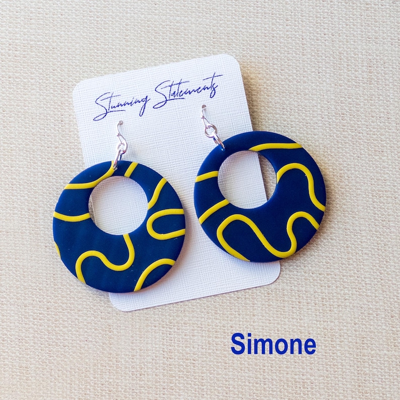K&ouml;nnte beinhalten: Ein Paar kreisf&ouml;rmige Ohrh&auml;nger mit dunkelblauer Basis und gelbem Wellenlinienmuster. Die Ohrringe sind an einer wei&szlig;en Pr&auml;sentationskarte befestigt, auf der der Text Stunning Statements und Simone in blauer Schrift aufgedruckt ist.