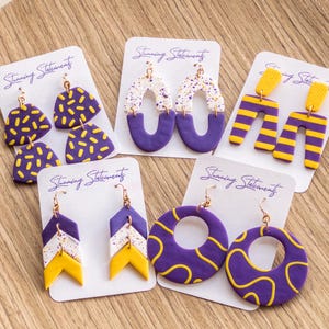 May include: Five pairs of polymer clay earrings displayed on white cards labeled Stunning Statements. Designs include purple with yellow dashes, white with purple and gold speckles, yellow and purple stripes, chevron patterns, and purple with yellow swirls.