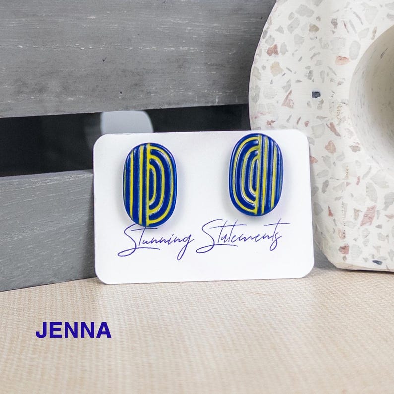 K&ouml;nnte beinhalten: Ein Paar ovale Ohrstecker mit einer tiefblauen Basis und gelben vertikalen sowie geschwungenen Linienmustern. Die Ohrringe sind auf einer wei&szlig;en Karte befestigt, auf der der Text Stunning Statements und der Name JENNA aufgedruckt sind.