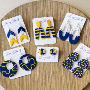 K&ouml;nnte beinhalten: Sechs Paar Ohrringe aus Polymer-Ton in Marineblau, Gelb und mit wei&szlig;en Sprenkelmustern, pr&auml;sentiert auf wei&szlig;en Karten mit der Aufschrift Stunning Statements. Die Designs umfassen Chevron-, Streifenbogen-, Oval-, Kreis-, Sechseck-Stecker- und F&auml;cherformen.