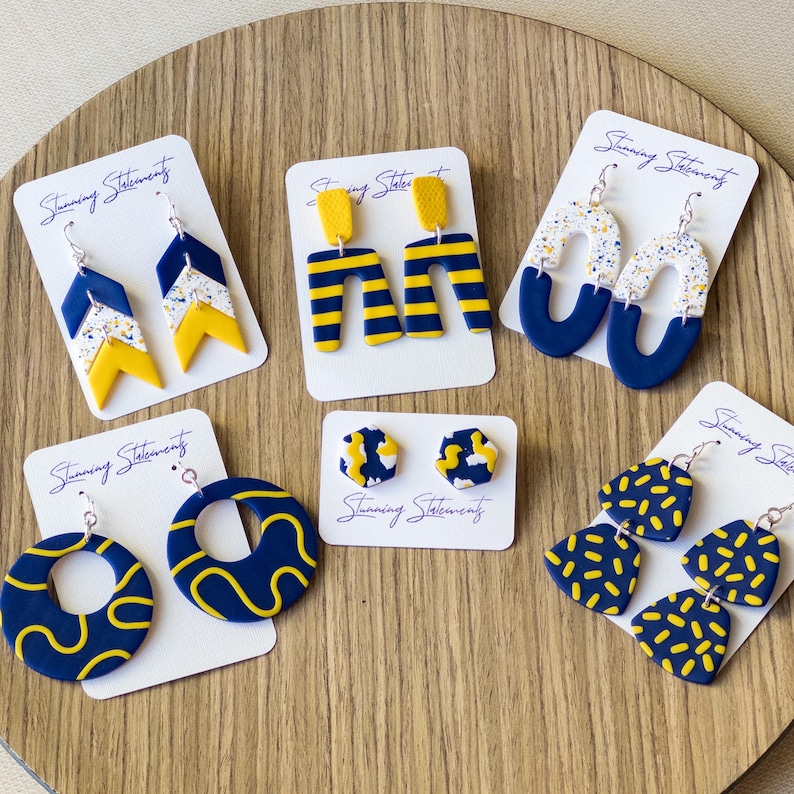 K&ouml;nnte beinhalten: Sechs Paar Ohrringe aus Polymer-Ton in Marineblau, Gelb und mit wei&szlig;en Sprenkelmustern, pr&auml;sentiert auf wei&szlig;en Karten mit der Aufschrift Stunning Statements. Die Designs umfassen Chevron-, Streifenbogen-, Oval-, Kreis-, Sechseck-Stecker- und F&auml;cherformen.