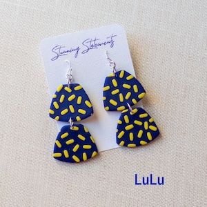 K&ouml;nnte beinhalten: Ein Paar H&auml;ngeohrringe bestehend aus zwei &uuml;bereinander angeordneten marineblauen Trapezformen mit gelbem Strichmuster. Die Ohrringe sind an einer wei&szlig;en Karte befestigt, auf der der Text Stunning Statements und LuLu in Blau steht.