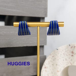 K&ouml;nnte beinhalten: Ein Paar dunkelblaue Huggie-Ohrringe mit d&uuml;nnen, vertikalen gelben Streifen. Die Ohrringe sind auf einem goldfarbenen T-f&ouml;rmigen Metallst&auml;nder ausgestellt. Das Wort HUGGIES ist in blauer Schrift unten links im Bild zu sehen.