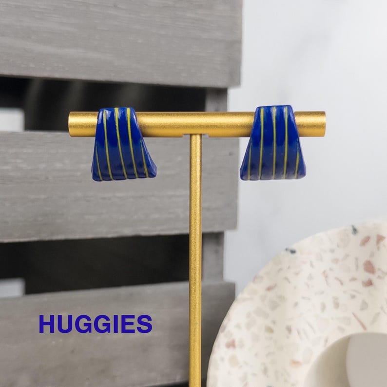 K&ouml;nnte beinhalten: Ein Paar dunkelblaue Huggie-Ohrringe mit d&uuml;nnen, vertikalen gelben Streifen. Die Ohrringe sind auf einem goldfarbenen T-f&ouml;rmigen Metallst&auml;nder ausgestellt. Das Wort HUGGIES ist in blauer Schrift unten links im Bild zu sehen.