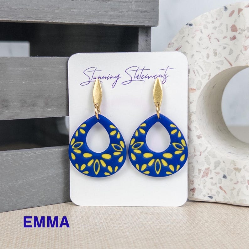 K&ouml;nnte beinhalten: Ein Paar tropfenf&ouml;rmige Ohrringe mit einer leuchtend blauen Basis und einem gelben Blumenmuster. Jeder Ohrring ist an einem goldfarbenen Metallstecker befestigt. Die Ohrringe sind auf einer wei&szlig;en Karte mit der Aufschrift Stunning Statements.