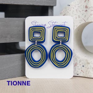 K&ouml;nnte beinhalten: Ein Paar Ohrh&auml;nger auf einer wei&szlig;en Schmuckkarte mit der Aufschrift Stunning Statements. Jeder Ohrring besteht aus einem blauen Quadrat mit gelben konzentrischen Linien, das mit einem blauen Kreis mit gelben konzentrischen Linien verbunden ist. Der Text TIONNE ist sichtbar.