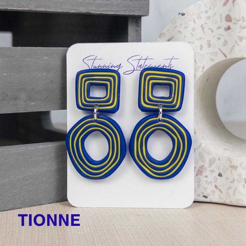 K&ouml;nnte beinhalten: Ein Paar Ohrh&auml;nger auf einer wei&szlig;en Schmuckkarte mit der Aufschrift Stunning Statements. Jeder Ohrring besteht aus einem blauen Quadrat mit gelben konzentrischen Linien, das mit einem blauen Kreis mit gelben konzentrischen Linien verbunden ist. Der Text TIONNE ist sichtbar.