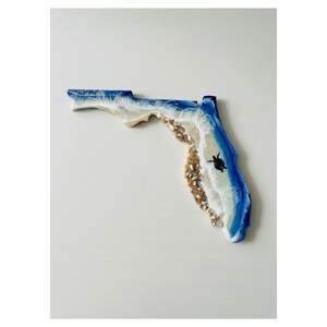 May include: A Florida-shaped resin art piece with a coastal theme. The design features blue and white resin representing the ocean, a sandy beach area with shells, and a black sea turtle. The artwork is a decorative item.