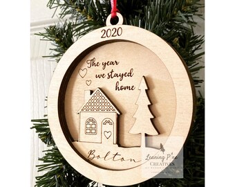 May include: A wooden Christmas ornament with a house and tree design. The ornament is engraved with the text "The year we stayed home" and the year "2020". The ornament also includes the name "Bolton" and the text "Leaning Pine Creatives".