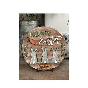 May include: A round wooden sign with the words "Hoppy Easter" in green and white lettering. Three grey bunny silhouettes are at the bottom of the sign, with a carrot and green leaves on the top. The sign is on a wooden stand with the name "Blakelee" written on it.