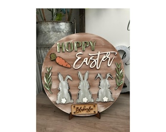 May include: A round wooden sign with the words "Hoppy Easter" in green and white lettering. Three grey bunny silhouettes are at the bottom of the sign, with a carrot and green leaves on the top. The sign is on a wooden stand with the name "Blakelee" written on it.