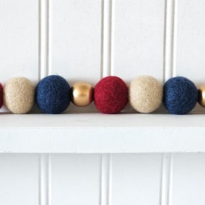 May include: A decorative garland featuring alternating red, beige, and navy blue felt balls, interspersed with gold-colored spheres. The garland is displayed against a white background, creating a festive and colorful accent.