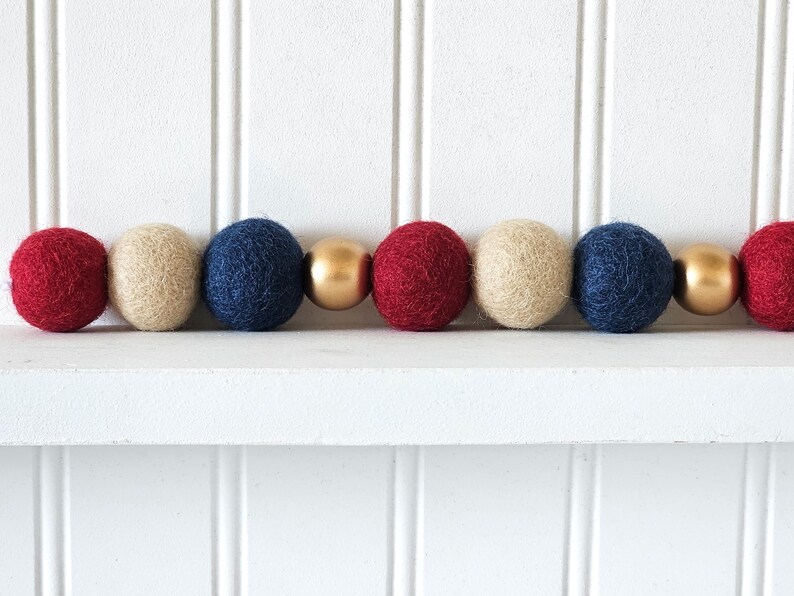 May include: A decorative garland featuring alternating red, beige, and navy blue felt balls, interspersed with gold-colored spheres. The garland is displayed against a white background, creating a festive and colorful accent.