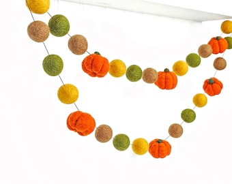 Puede incluir: Una guirnalda decorativa con bolas de lana fieltrada en tonos amarillo, verde oliva y marrón, intercaladas con pequeñas calabazas de fieltro naranja que tienen tallos verdes. Los elementos están unidos por un cordón fino.