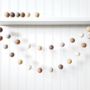 Puede incluir: Una guirnalda decorativa de bolas de fieltro en tonos neutros de blanco, beige y marr&oacute;n. La guirnalda est&aacute; formada por m&uacute;ltiples bolas de fieltro, creando un acento decorativo. La guirnalda se exhibe en una estanter&iacute;a blanca contra una pared blanca con paneles.