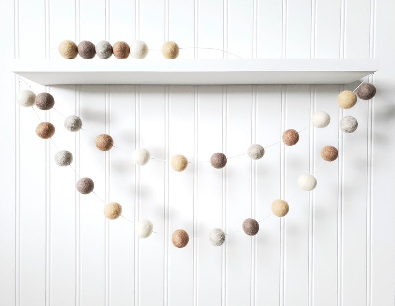 Puede incluir: Una guirnalda decorativa de bolas de fieltro en tonos neutros de blanco, beige y marr&oacute;n. La guirnalda est&aacute; formada por m&uacute;ltiples bolas de fieltro, creando un acento decorativo. La guirnalda se exhibe en una estanter&iacute;a blanca contra una pared blanca con paneles.