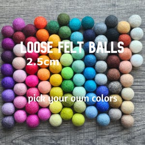 May include: A collection of colorful, 2.5cm felt balls arranged on a wooden surface. The balls are in various shades of purple, pink, orange, yellow, green, blue, gray, brown, and white. Text on the image reads "LOOSE FELT BALLS 2.5cm pick your own colors."