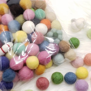 May include: A clear plastic bag filled with a variety of colorful felt balls. The balls are in various shades of pink, blue, green, yellow, orange, and red. There are also a few white felt balls in the bag.