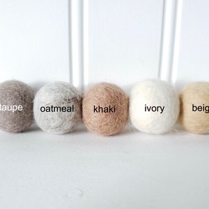 Puede incluir: Cinco bolas de fieltro en fila, cada una de un color neutro diferente. Los colores son topo, avena, caqui, marfil y beige. Cada bola tiene su nombre de color.