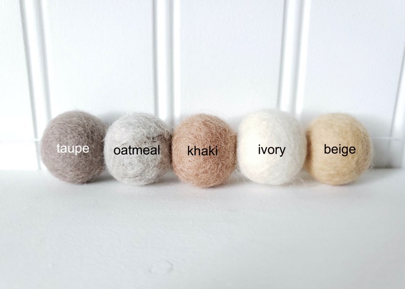 Puede incluir: Cinco bolas de fieltro en fila, cada una de un color neutro diferente. Los colores son topo, avena, caqui, marfil y beige. Cada bola tiene su nombre de color.