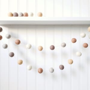 Puede incluir: Guirnalda decorativa de bolas de fieltro en tonos neutros, incluyendo blanco, beige, gris y marr&oacute;n. La guirnalda est&aacute; ensartada en un fino cord&oacute;n blanco y cuelga en una suave curva sobre un fondo blanco. Bolas de fieltro adicionales en una estanter&iacute;a.
