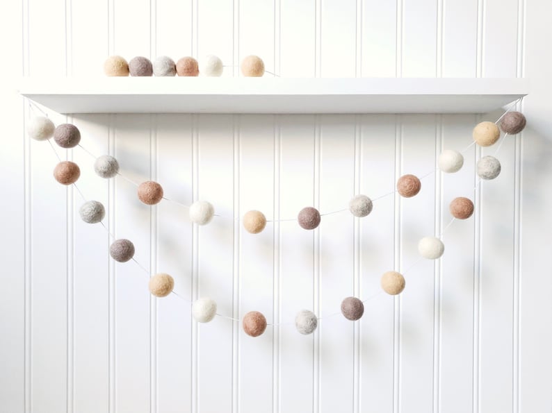 Puede incluir: Guirnalda decorativa de bolas de fieltro en tonos neutros, incluyendo blanco, beige, gris y marr&oacute;n. La guirnalda est&aacute; ensartada en un fino cord&oacute;n blanco y cuelga en una suave curva sobre un fondo blanco. Bolas de fieltro adicionales en una estanter&iacute;a.
