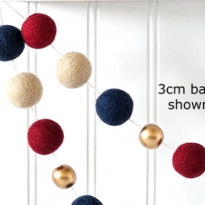 May include: A garland of felt balls in navy blue, red, cream, and gold. The text "3cm balls shown" is visible. The balls are strung together on a white string, creating a festive decoration.