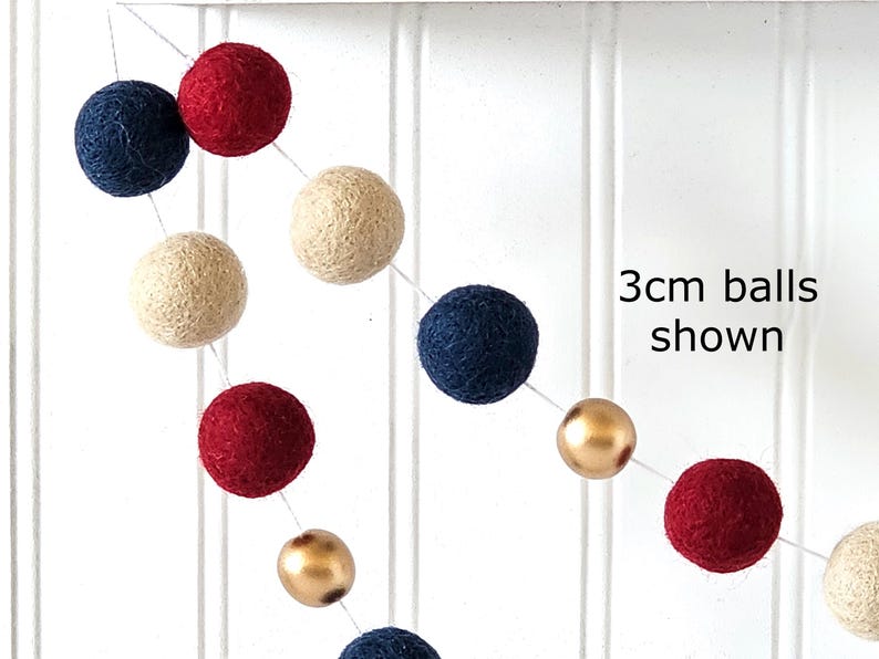 May include: A garland of felt balls in navy blue, red, cream, and gold. The text "3cm balls shown" is visible. The balls are strung together on a white string, creating a festive decoration.