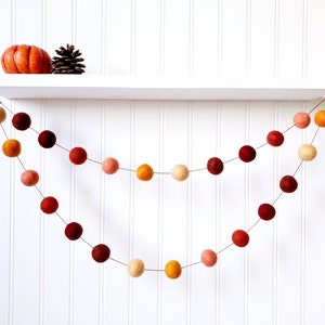 May include: A garland of felt balls in shades of orange, red, and cream hangs from a white shelf. The garland is made of two strands of felt balls, with the top strand being slightly longer than the bottom strand.