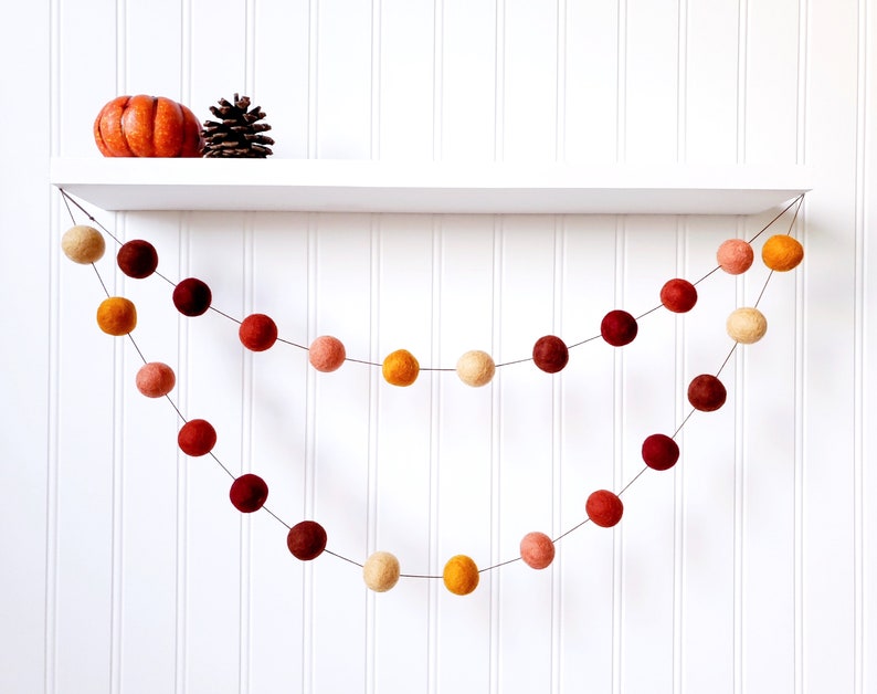 May include: A garland of felt balls in shades of orange, red, and cream hangs from a white shelf. The garland is made of two strands of felt balls, with the top strand being slightly longer than the bottom strand.