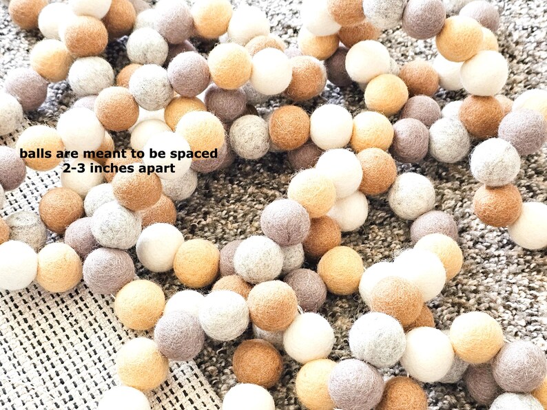 Puede incluir: Una guirnalda de bolas de fieltro en varios colores neutros, incluyendo blanco, beige, gris y marr&oacute;n. El texto en la imagen dice: "las bolas deben espaciarse 5-7,6 cm". Las bolas est&aacute;n ensartadas, creando un elemento decorativo.