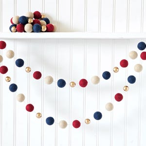 May include: A festive garland featuring red, white, and navy blue felt balls interspersed with gold metallic beads. A collection of similar felt balls rests on a white shelf above the garland. The background is a white vertical shiplap wall.