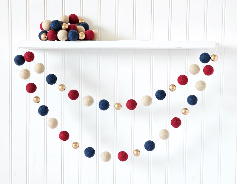 May include: A festive garland featuring red, white, and navy blue felt balls interspersed with gold metallic beads. A collection of similar felt balls rests on a white shelf above the garland. The background is a white vertical shiplap wall.