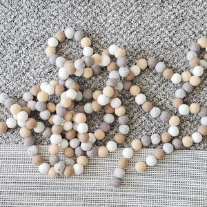 Puede incluir: Una guirnalda decorativa compuesta por bolas de fieltro en tonos blanco, beige y gris. Las bolas est&aacute;n ensartadas para crear una hebra larga y flexible, adecuada para la decoraci&oacute;n del hogar u ocasiones festivas.