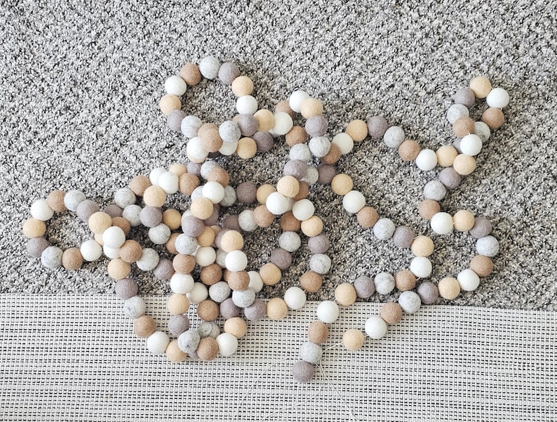 Puede incluir: Una guirnalda decorativa compuesta por bolas de fieltro en tonos blanco, beige y gris. Las bolas est&aacute;n ensartadas para crear una hebra larga y flexible, adecuada para la decoraci&oacute;n del hogar u ocasiones festivas.