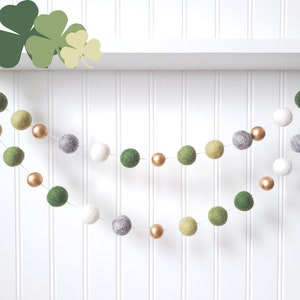 May include: A green, gray, and gold felt ball garland with a white background. The garland is made of felt balls in various sizes and colors, including green, gray, and gold. The garland is strung on a white string and is hanging from a white shelf.