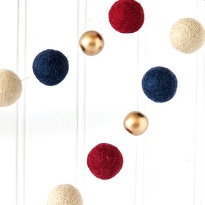 May include: A garland of felt balls in various colors, including cream, navy blue, red, and gold. The balls are strung together, creating a decorative element. The background is a white surface with vertical lines.