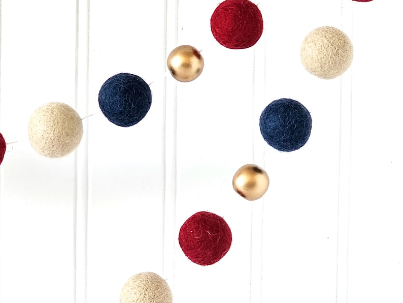 May include: A garland of felt balls in various colors, including cream, navy blue, red, and gold. The balls are strung together, creating a decorative element. The background is a white surface with vertical lines.