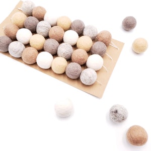 Puede incluir: Un conjunto de bolas de fieltro en varios colores neutros, incluyendo blanco, beige y marr&oacute;n, dispuestas sobre una tarjeta marr&oacute;n. Algunas bolas est&aacute;n ensartadas, otras dispersas. Las bolas son suaves y texturizadas.