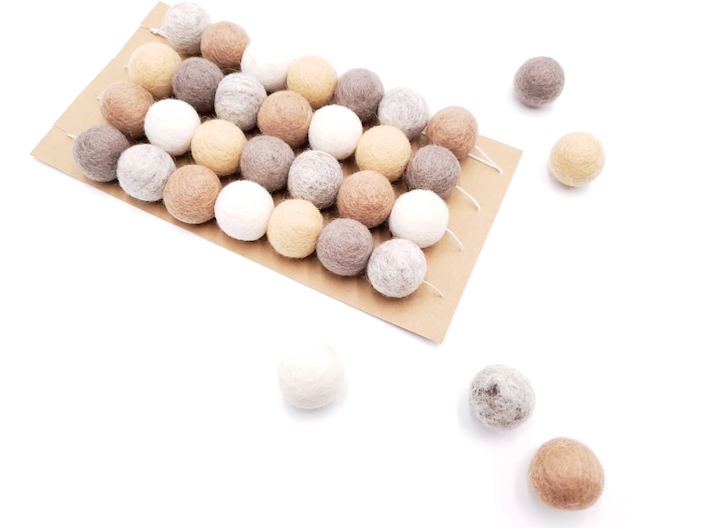 Puede incluir: Un conjunto de bolas de fieltro en varios colores neutros, incluyendo blanco, beige y marr&oacute;n, dispuestas sobre una tarjeta marr&oacute;n. Algunas bolas est&aacute;n ensartadas, otras dispersas. Las bolas son suaves y texturizadas.