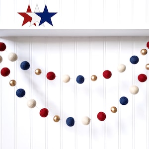 May include: A patriotic garland featuring red, white, and blue felt balls interspersed with gold beads. The garland is suspended against a white backdrop, with star-shaped decorations above. Perfect for festive decor.