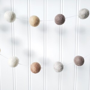 Puede incluir: Una guirnalda de bolas de fieltro en colores neutros, incluyendo blanco, beige, marr&oacute;n y gris. La guirnalda est&aacute; ensartada en un fino hilo blanco y cuelga sobre un fondo blanco con lamas verticales.