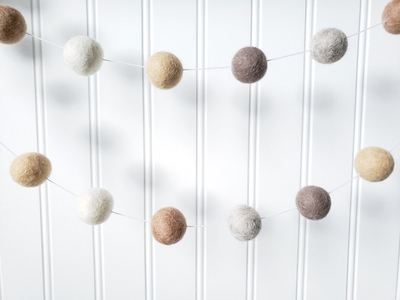 Puede incluir: Una guirnalda de bolas de fieltro en colores neutros, incluyendo blanco, beige, marr&oacute;n y gris. La guirnalda est&aacute; ensartada en un fino hilo blanco y cuelga sobre un fondo blanco con lamas verticales.