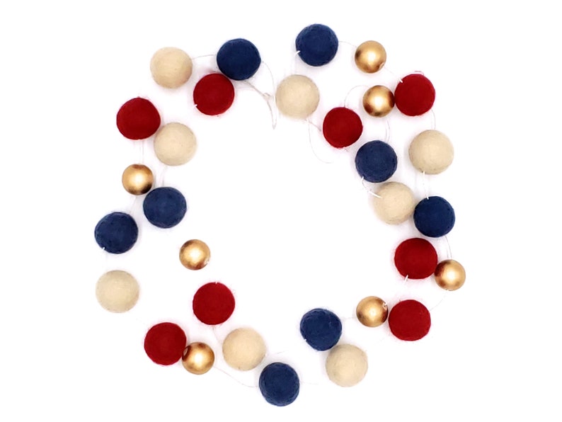 May include: A festive garland featuring alternating red, navy blue, cream-colored felt balls, and gold metallic spheres. The garland is strung on a thin white cord, creating a decorative accent for celebrations and home decor.